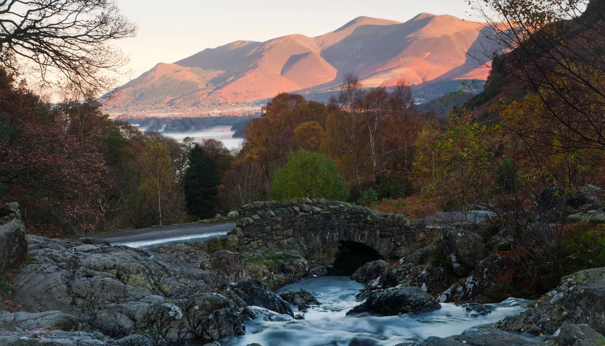 Stone bridge over a fast-flowing stream beneath sunlit mountains and autumn woodland in the Lake District.