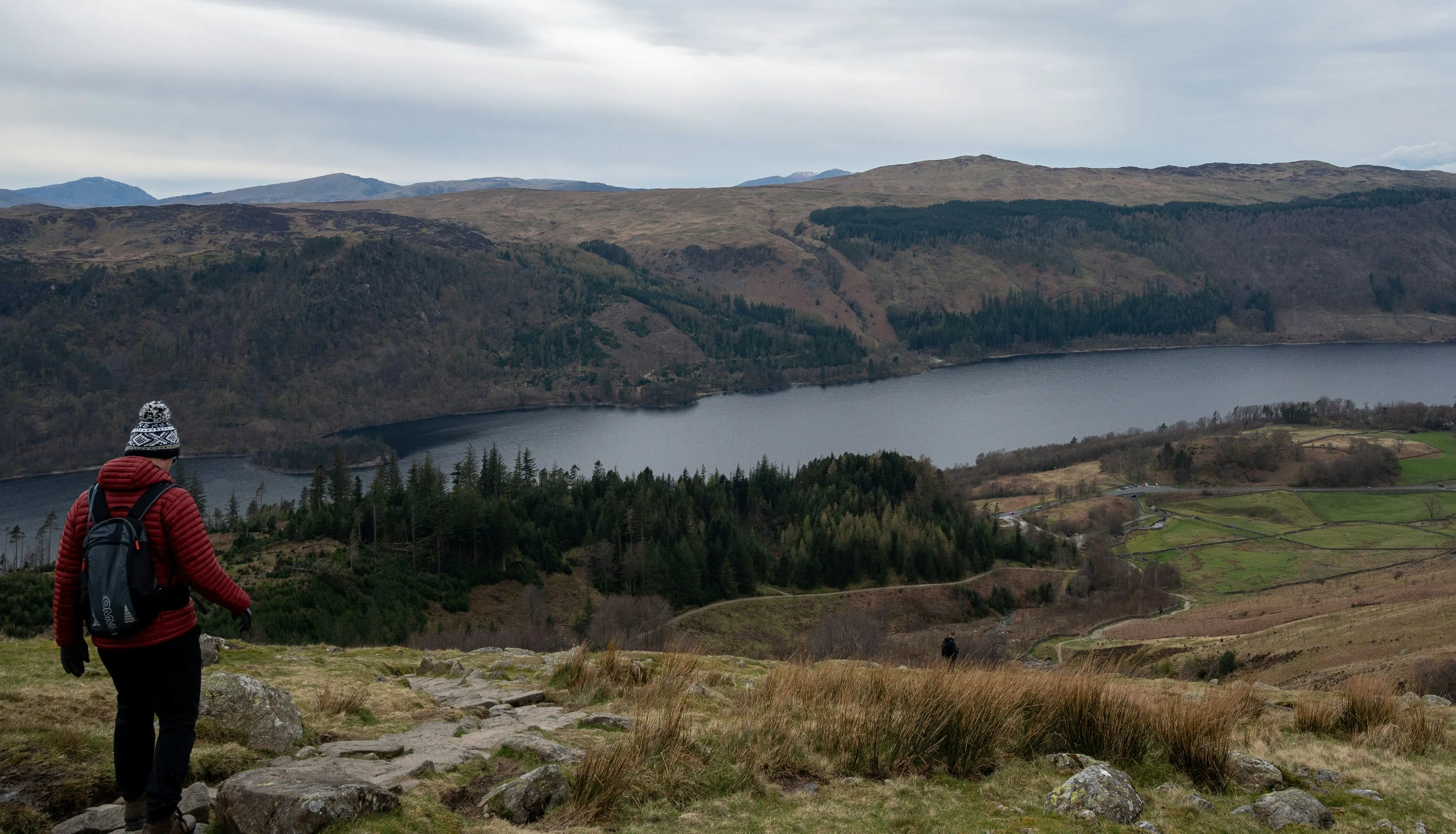 Hiker in a red jacket overlooking a lake, wooded hills and patchwork fields beneath an overcast sky.