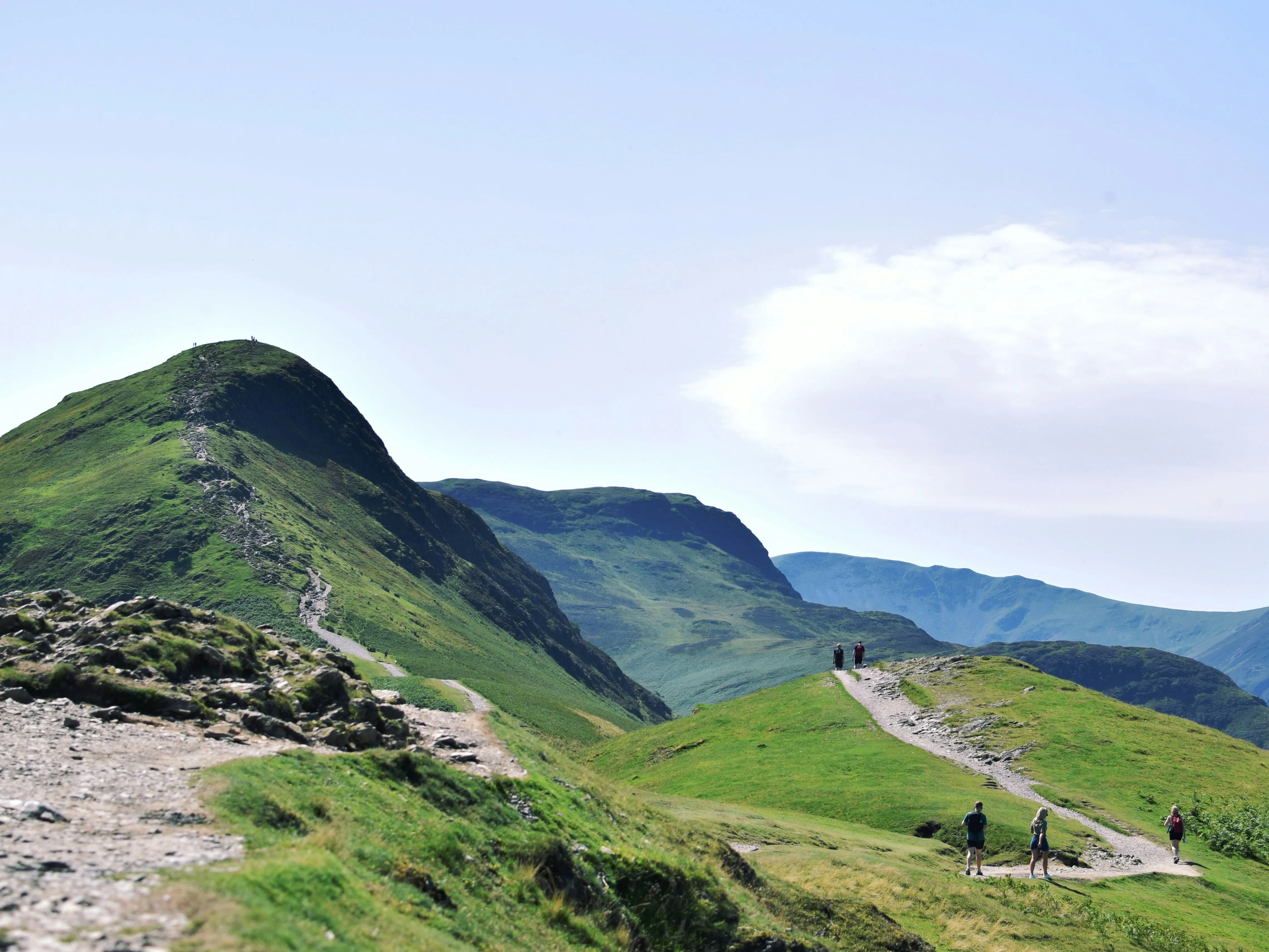 Hikers walking along a winding rocky trail through green mountain hills beneath a bright blue sky.