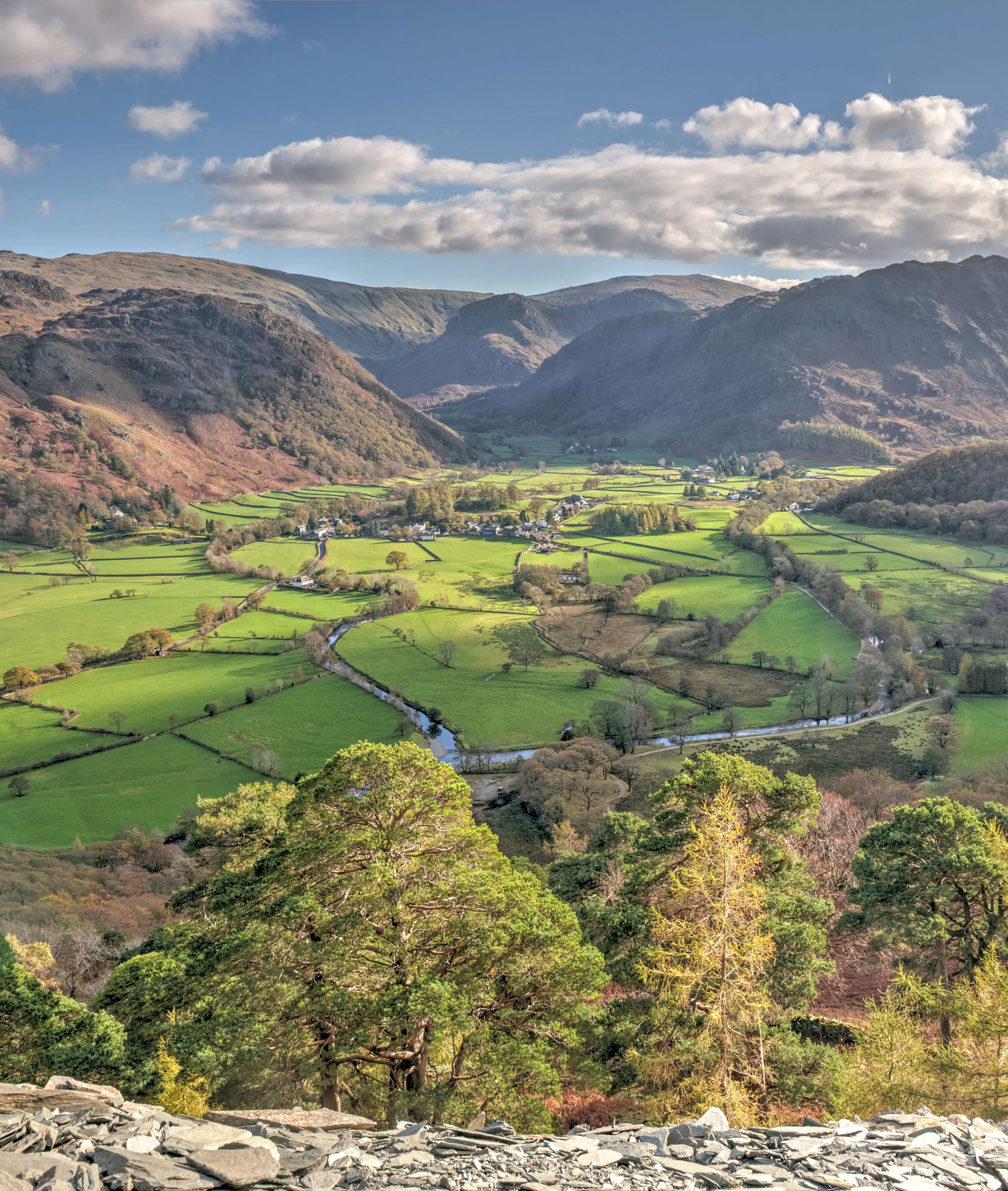 Sunlit view across Borrowdale Valley, with green patchwork fields, winding river, woodland and rugged Lake District fells beneath a cloud-filled blue sky.