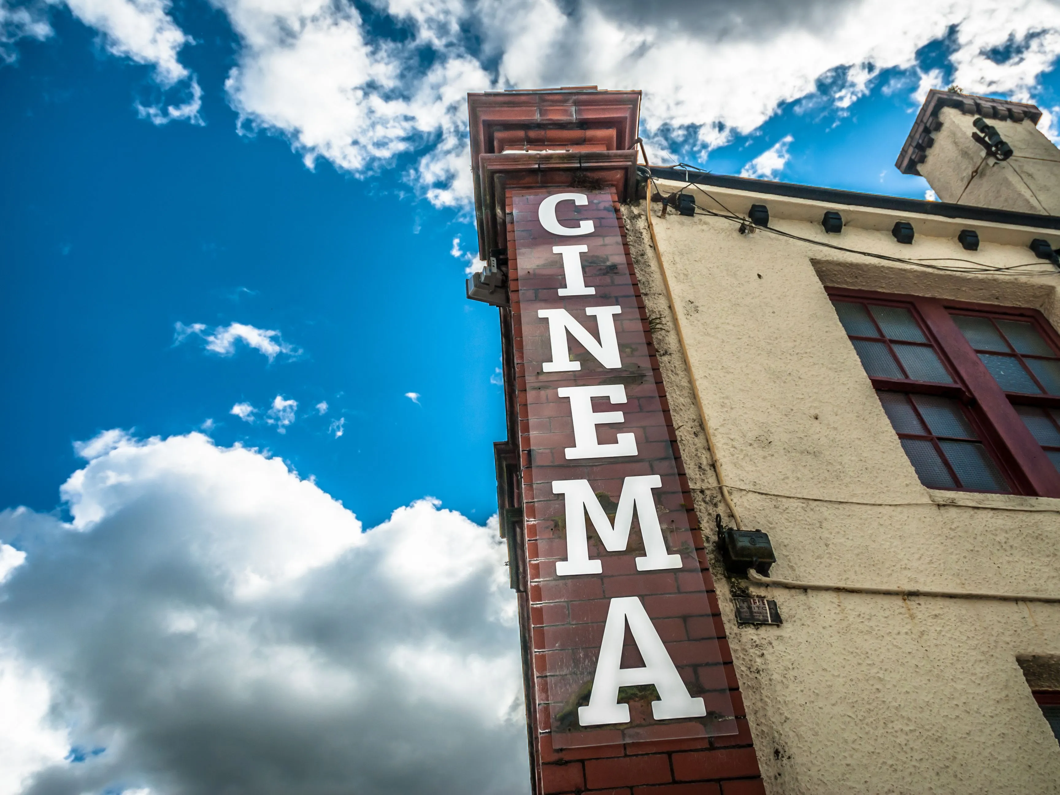 Low-angle view of a cinema sign on an old building against a vivid blue sky with clouds.