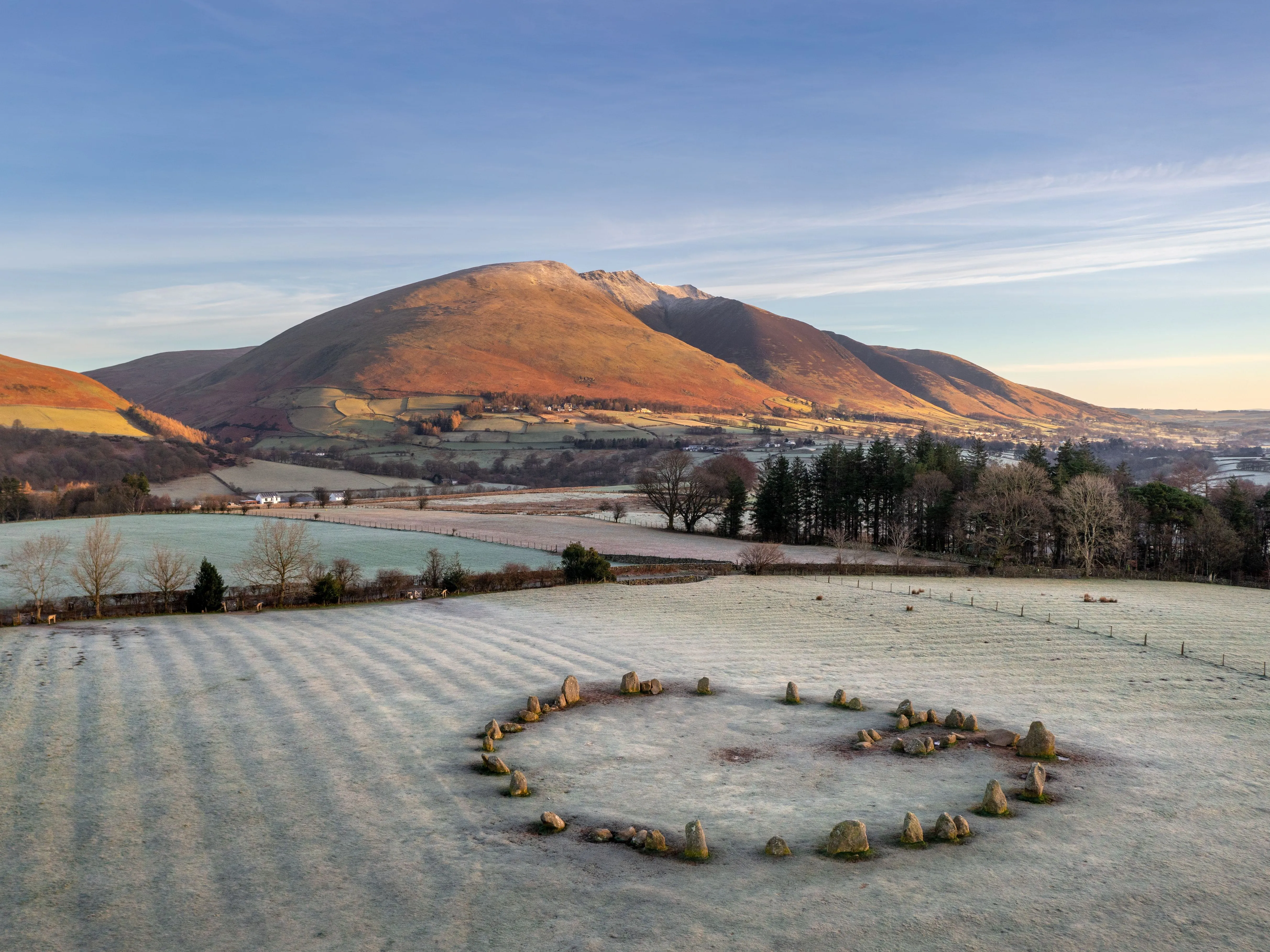 Ancient stone circle in a frost-covered field beneath sunlit mountains and a clear winter sky.