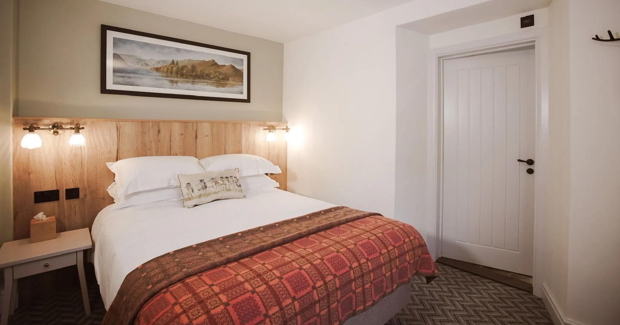 Cosy hotel bedroom with a double bed, wooden headboard, wall lights and patterned red throw.