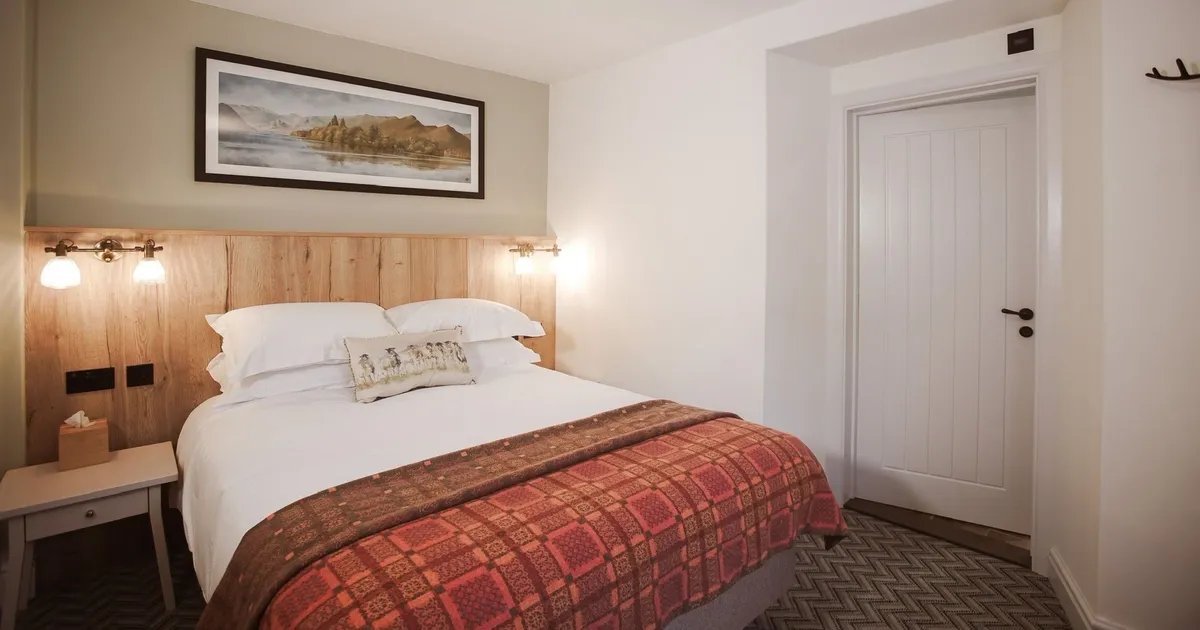 Cosy hotel bedroom with a double bed, wooden headboard, wall lights and patterned red throw.