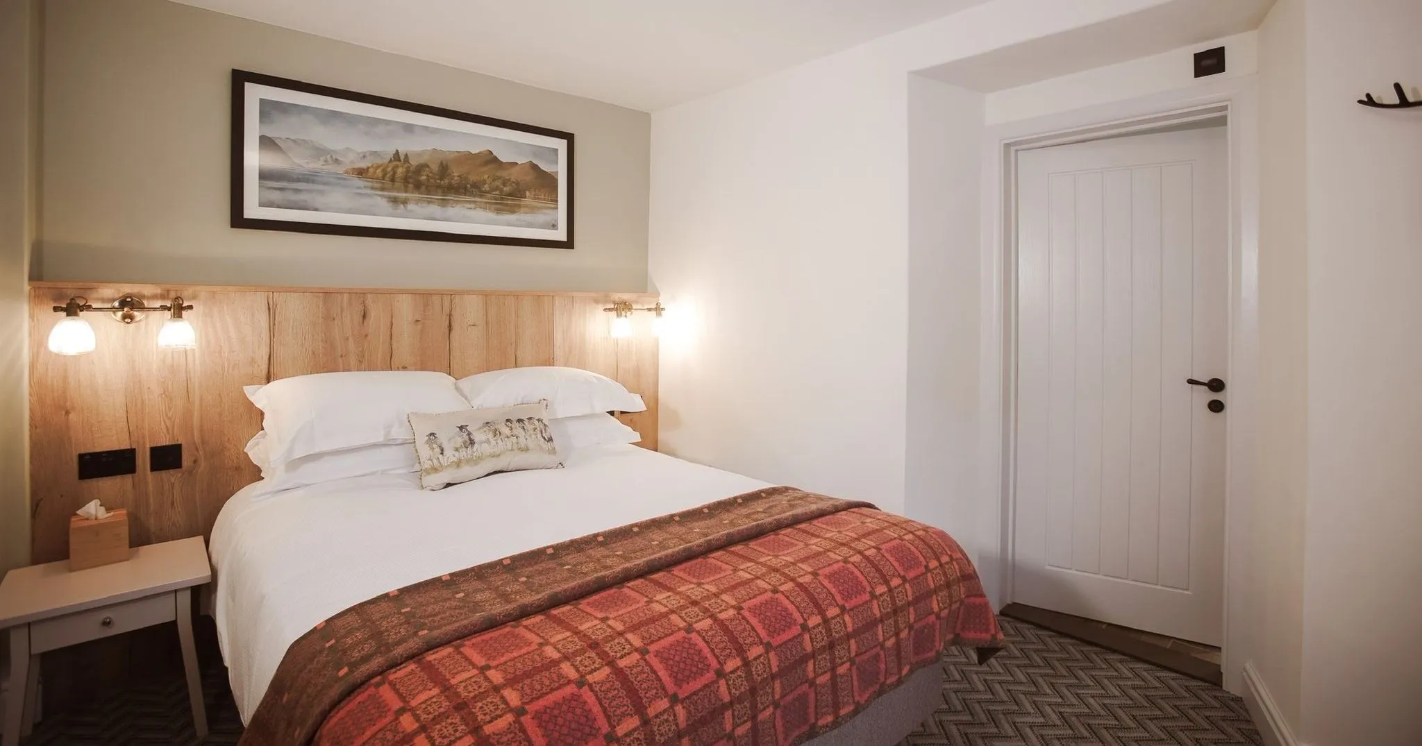 Cosy hotel bedroom with a double bed, wooden headboard, wall lights and patterned red throw.