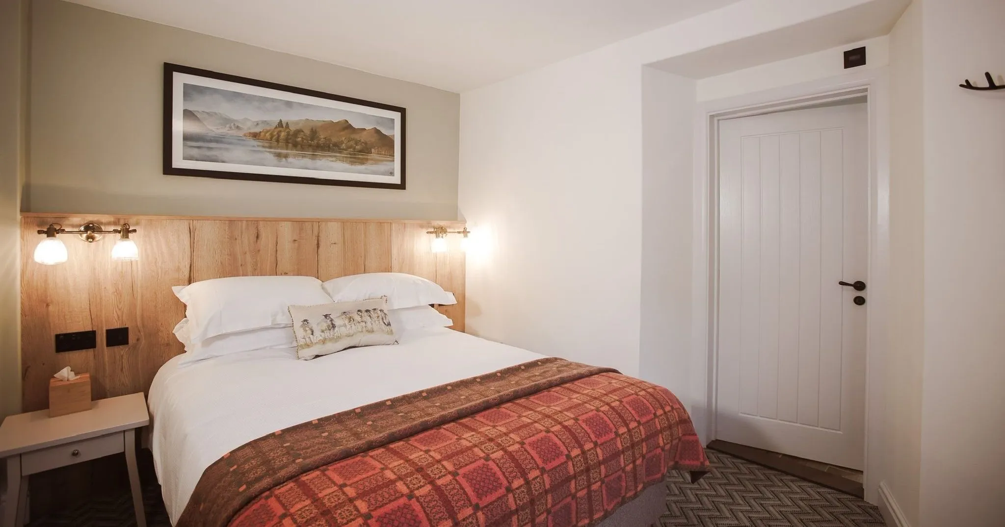 Cosy hotel bedroom with a double bed, wooden headboard, wall lights and patterned red throw.