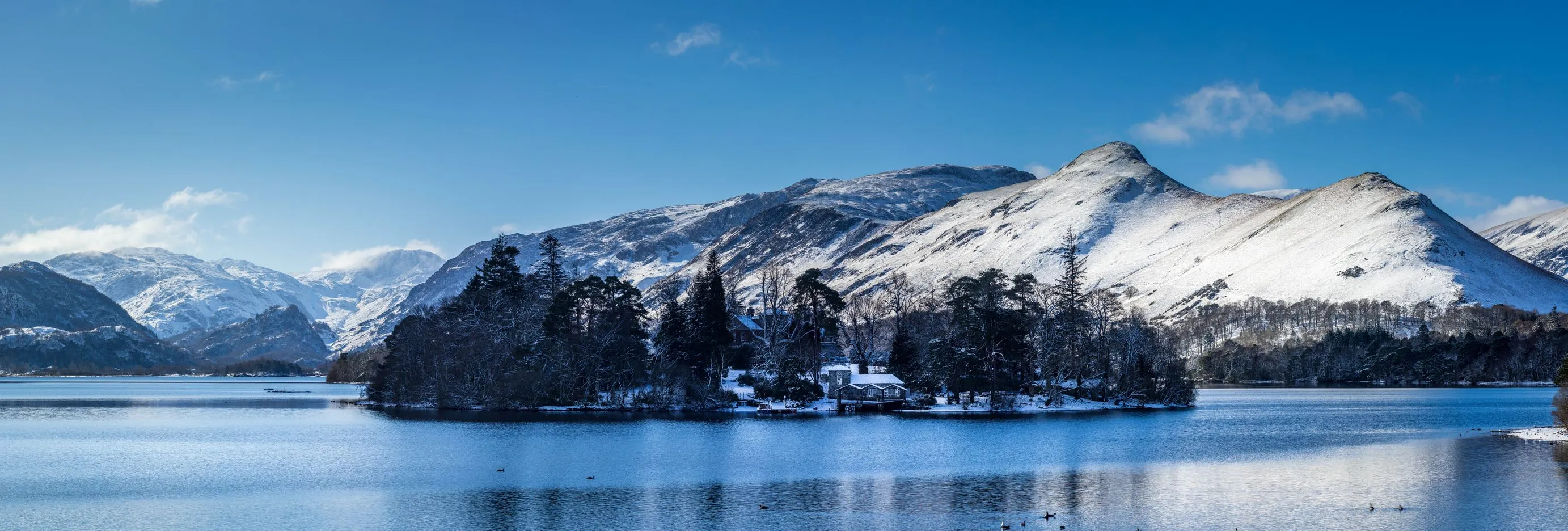 Snow-covered mountains rise behind a wooded island on a calm lake beneath a clear blue sky.