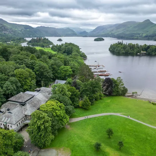Aerial view of a tranquil lake with wooded islands, boats, green parkland and mountains beneath cloudy skies.