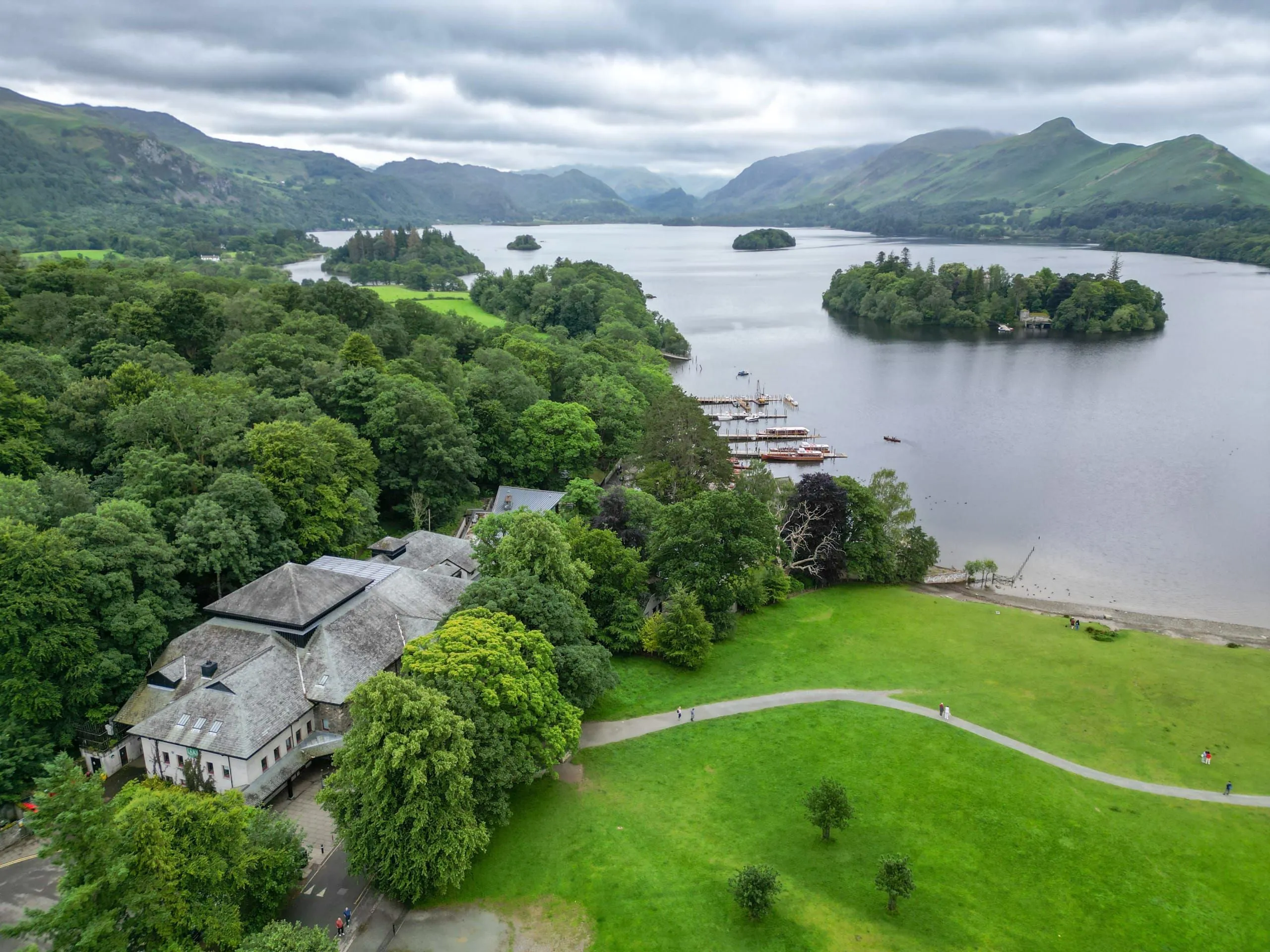 Aerial view of a tranquil lake with wooded islands, boats, green parkland and mountains beneath cloudy skies.