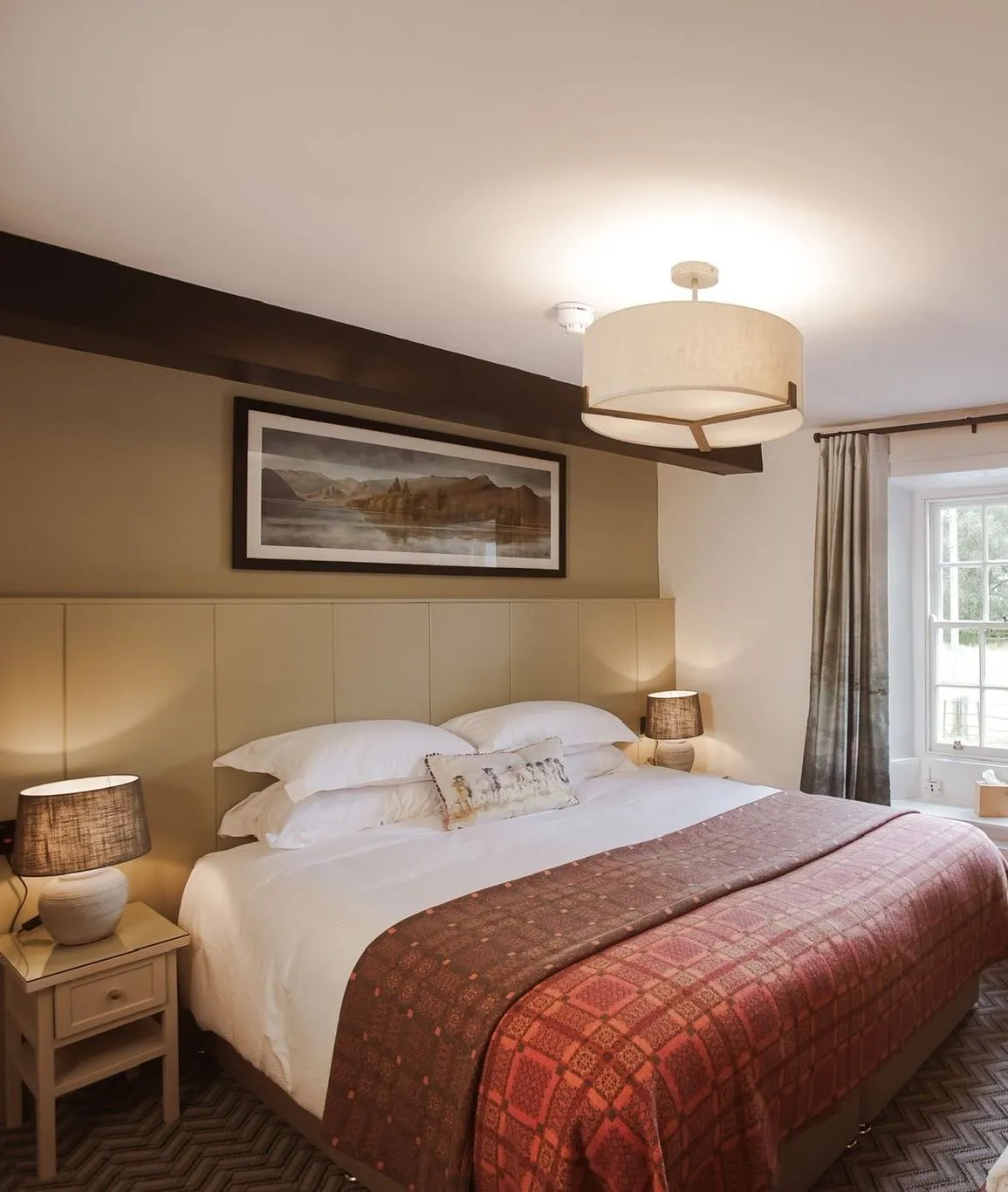 Cosy hotel bedroom with a double bed, patterned red throw, bedside lamps and a curtained window.