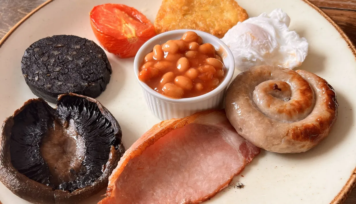 Full English breakfast plate with bacon, sausage, black pudding, baked beans, poached egg, hash brown, tomato and mushroom.