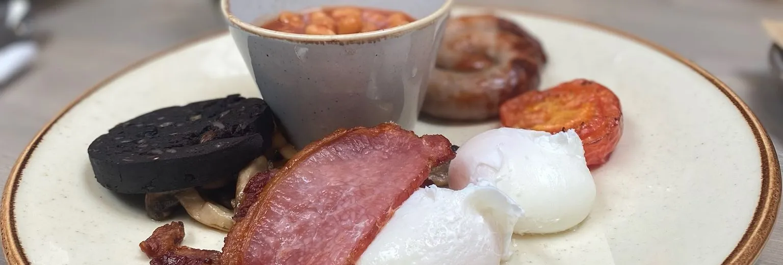 Cooked breakfast plate with bacon, poached eggs, black pudding, sausage, tomato and baked beans.