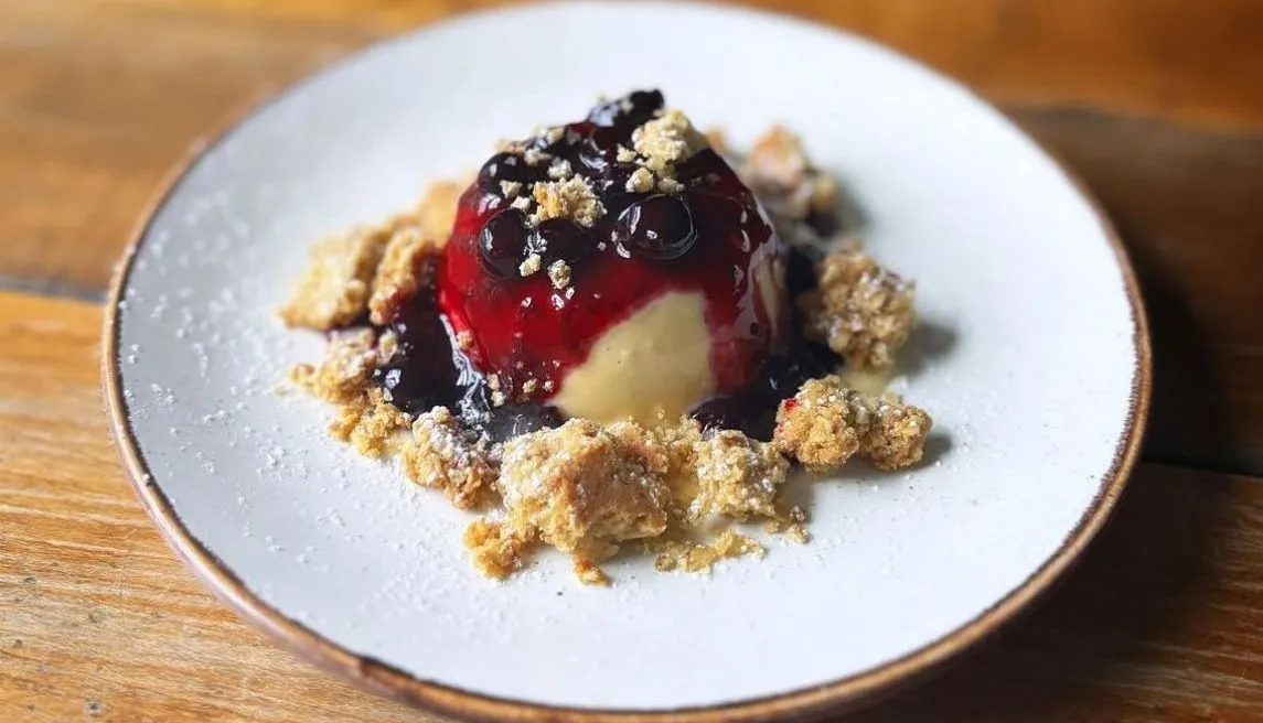 Vanilla panna cotta topped with dark berry compote and crumble on a white plate