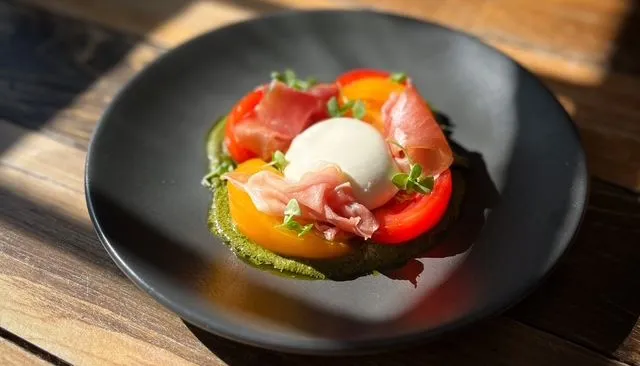 Black plate of heirloom tomatoes, burrata, prosciutto and green pesto on a sunlit wooden table