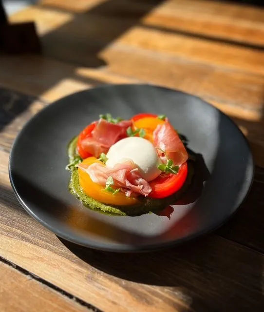 Black plate of heirloom tomatoes, burrata, prosciutto and green pesto on a sunlit wooden table
