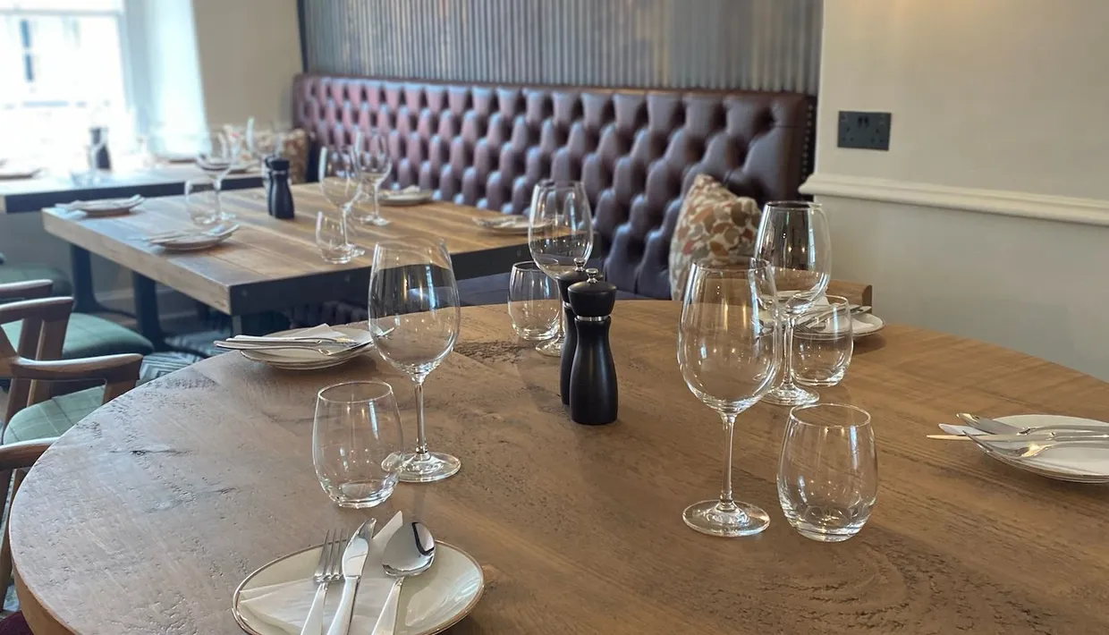 Cosy restaurant dining area with wooden tables set with wine glasses, plates and cutlery beside a tufted leather banquette.