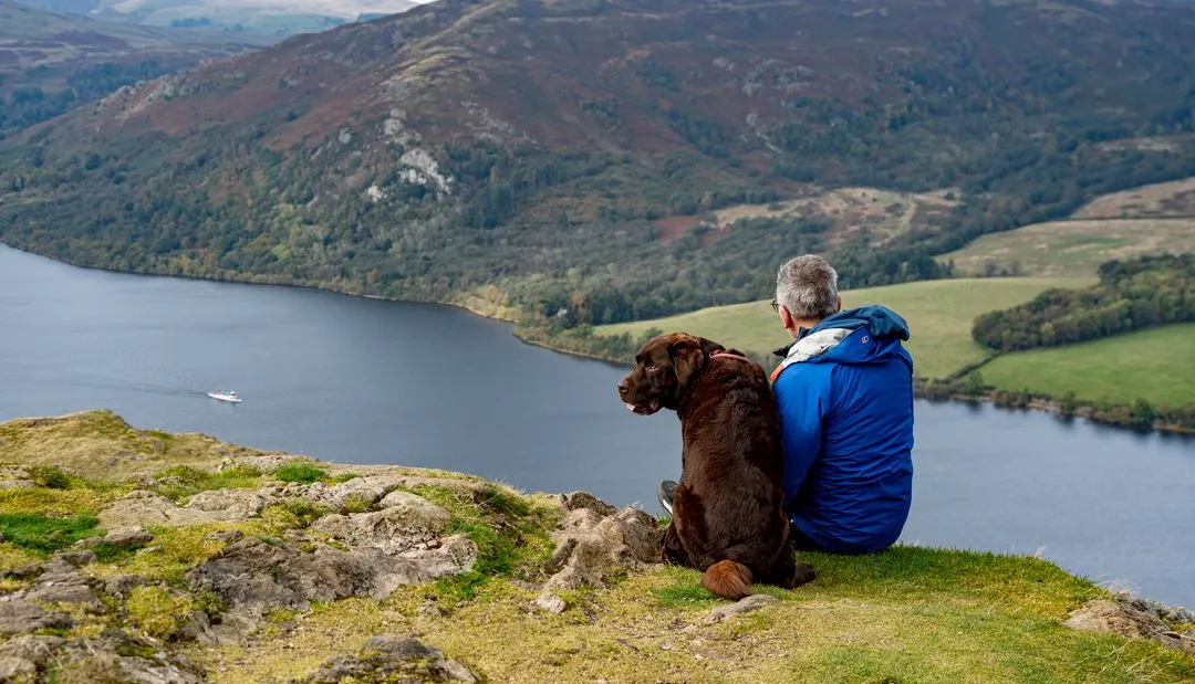 Hiker in a blue jacket sitting with a brown dog above Ullswater in the Lake District.
