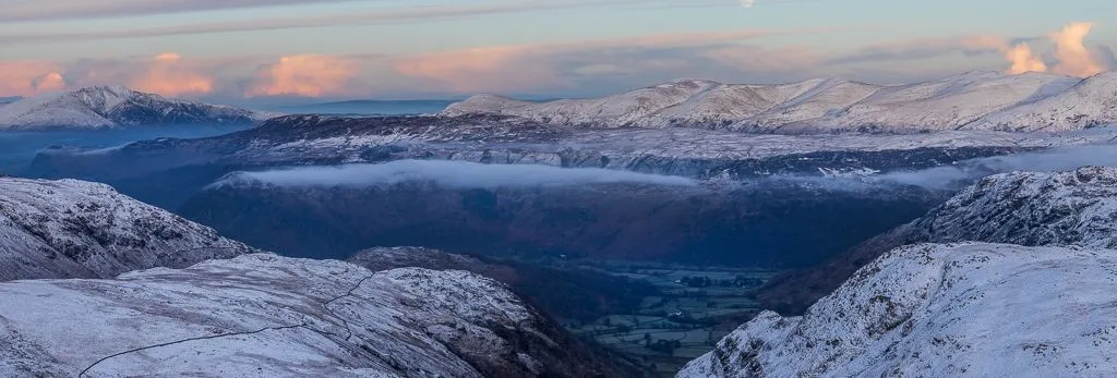 Snow-covered fells overlooking the Borrowdale Valley beneath layered clouds and a pale moon in winter.