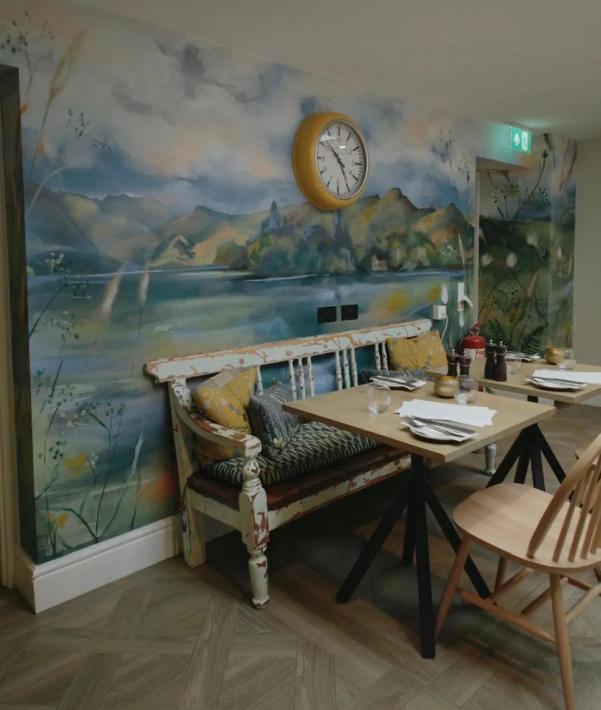 Cosy dining area with a painted mountain landscape mural, distressed bench, wooden table and wall clock.