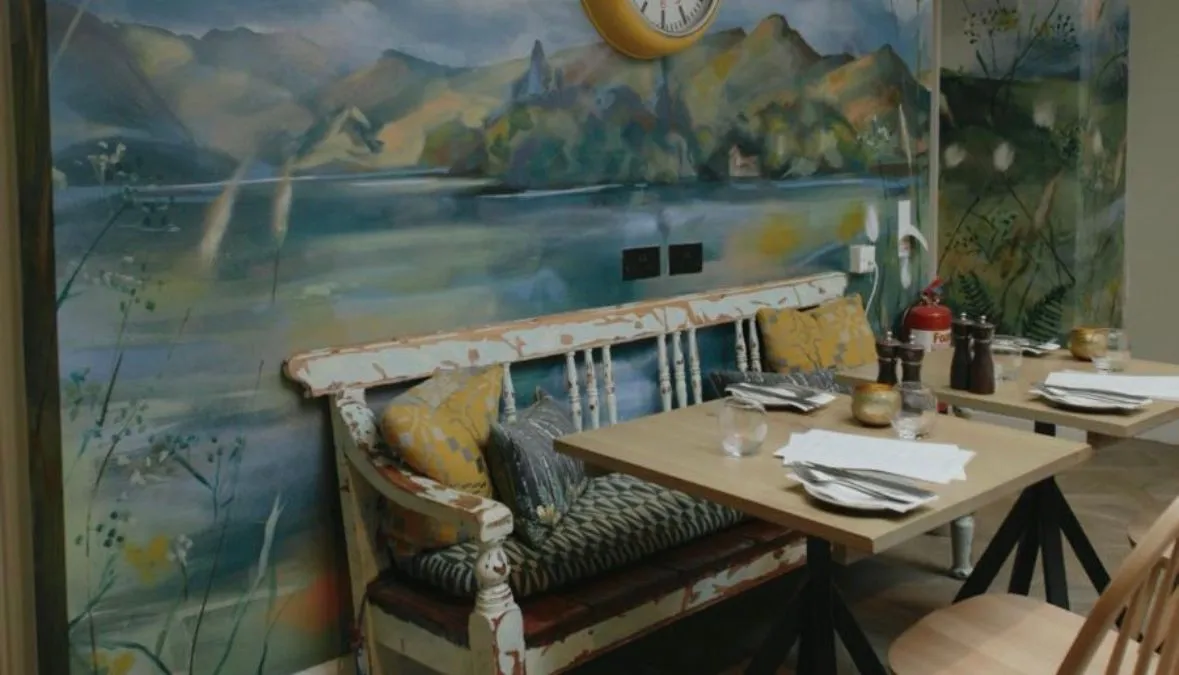 Cosy dining area with a painted mountain landscape mural, distressed bench, wooden table and wall clock.