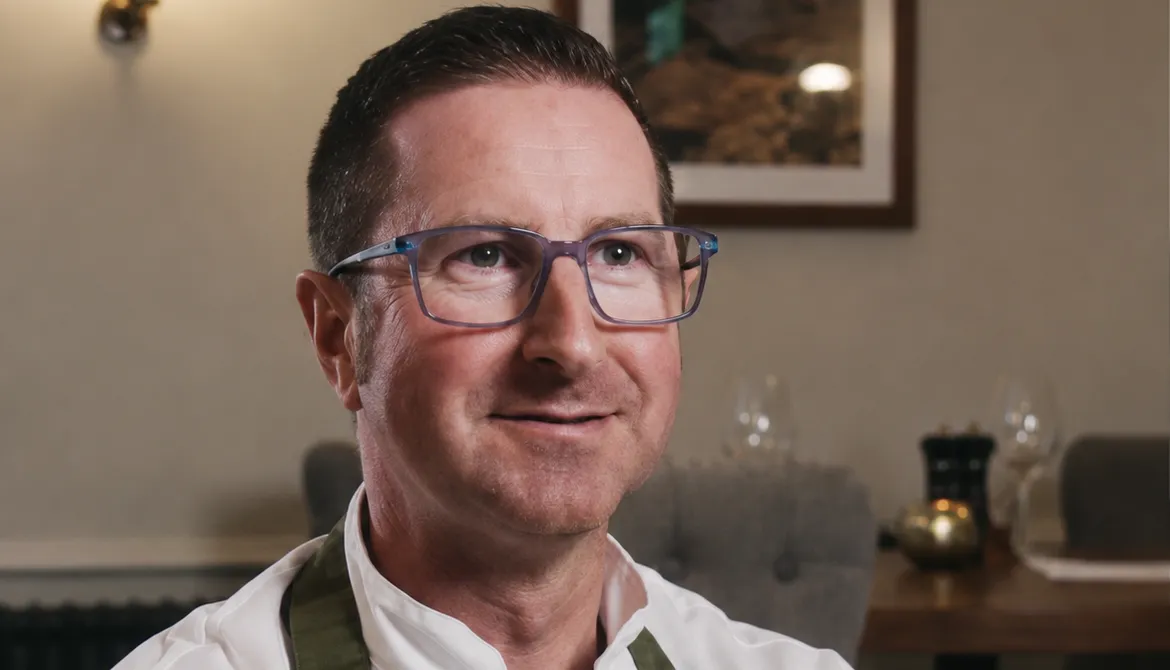 Portrait of David, a chef wearing glasses, a white chef's jacket and a green apron in a restaurant dining room.
