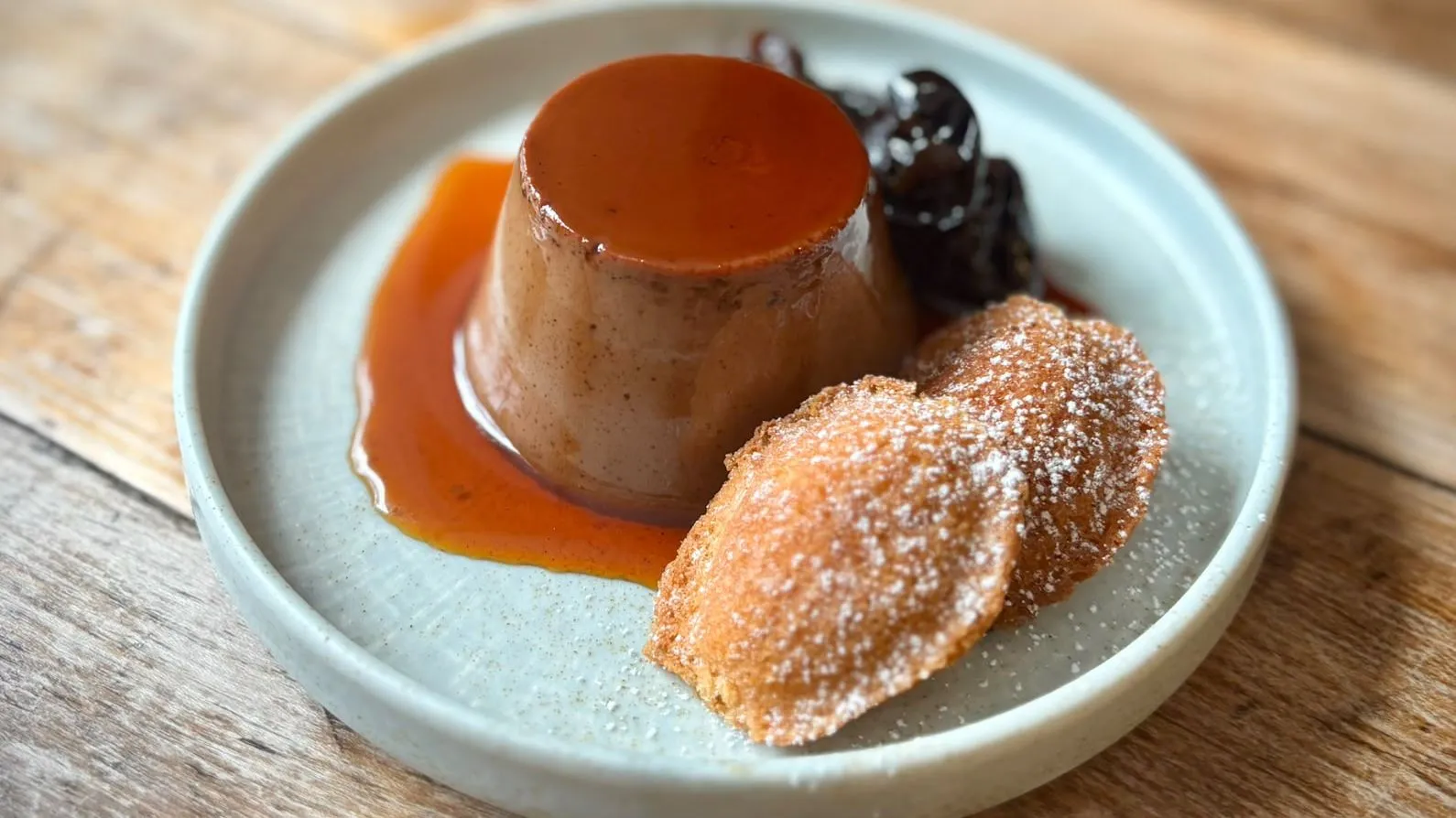 Caramel-topped pudding served with stewed dark fruit and a sugar-dusted churro on a ceramic plate