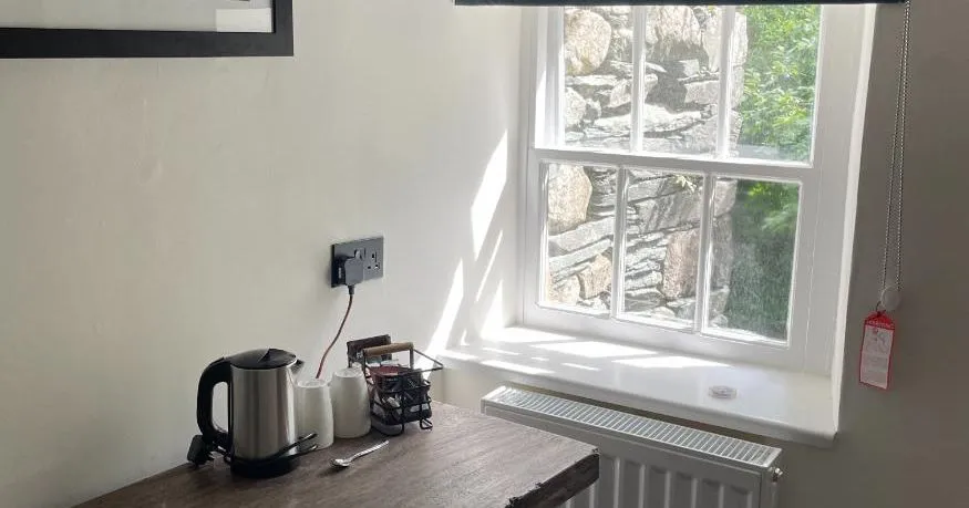 Cosy room corner with a wooden desk, kettle, grey Roman blind and sunlit sash window.