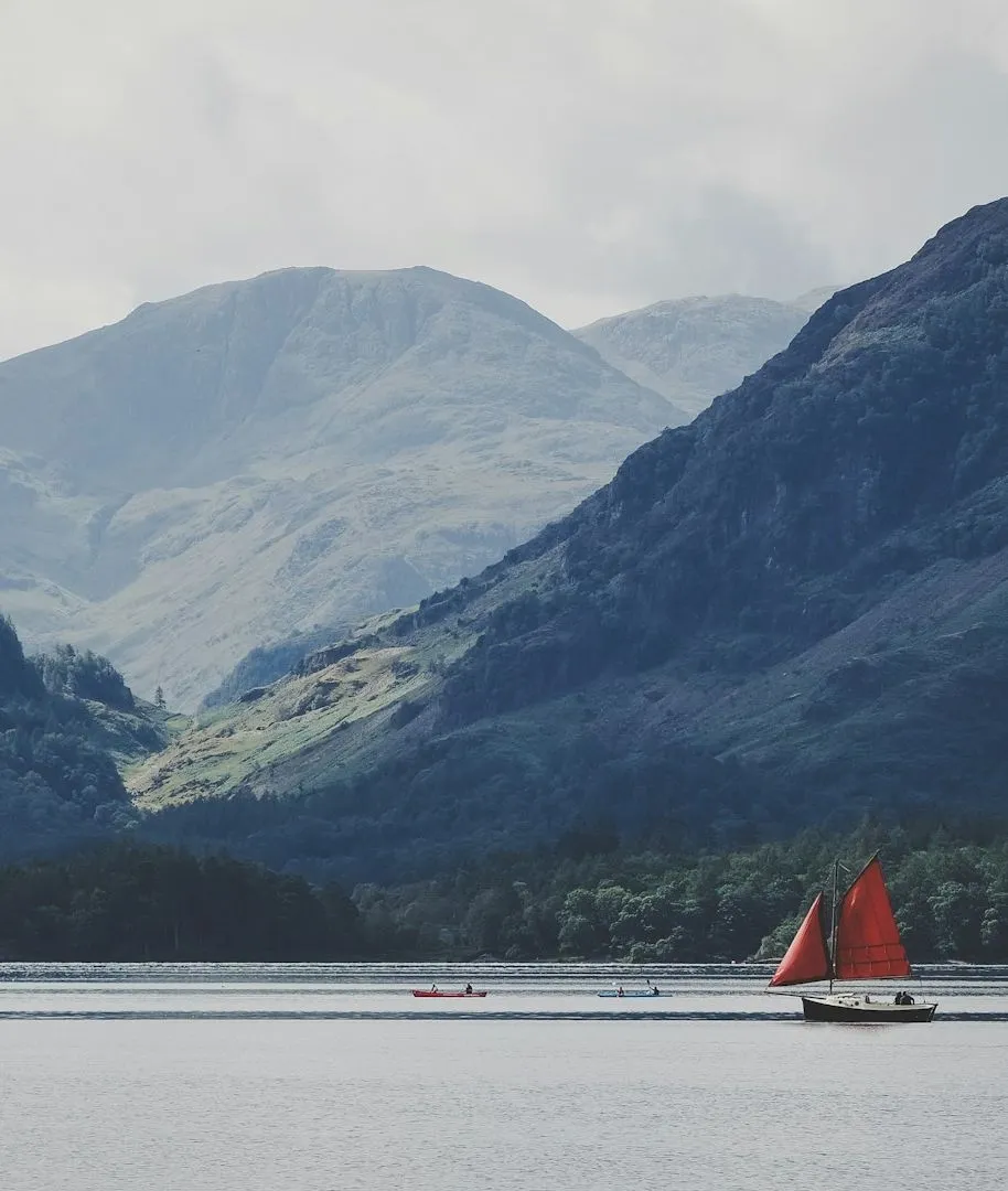 Sailing boat with bright red sails on Derwentwater beneath misty Lake District fells.
