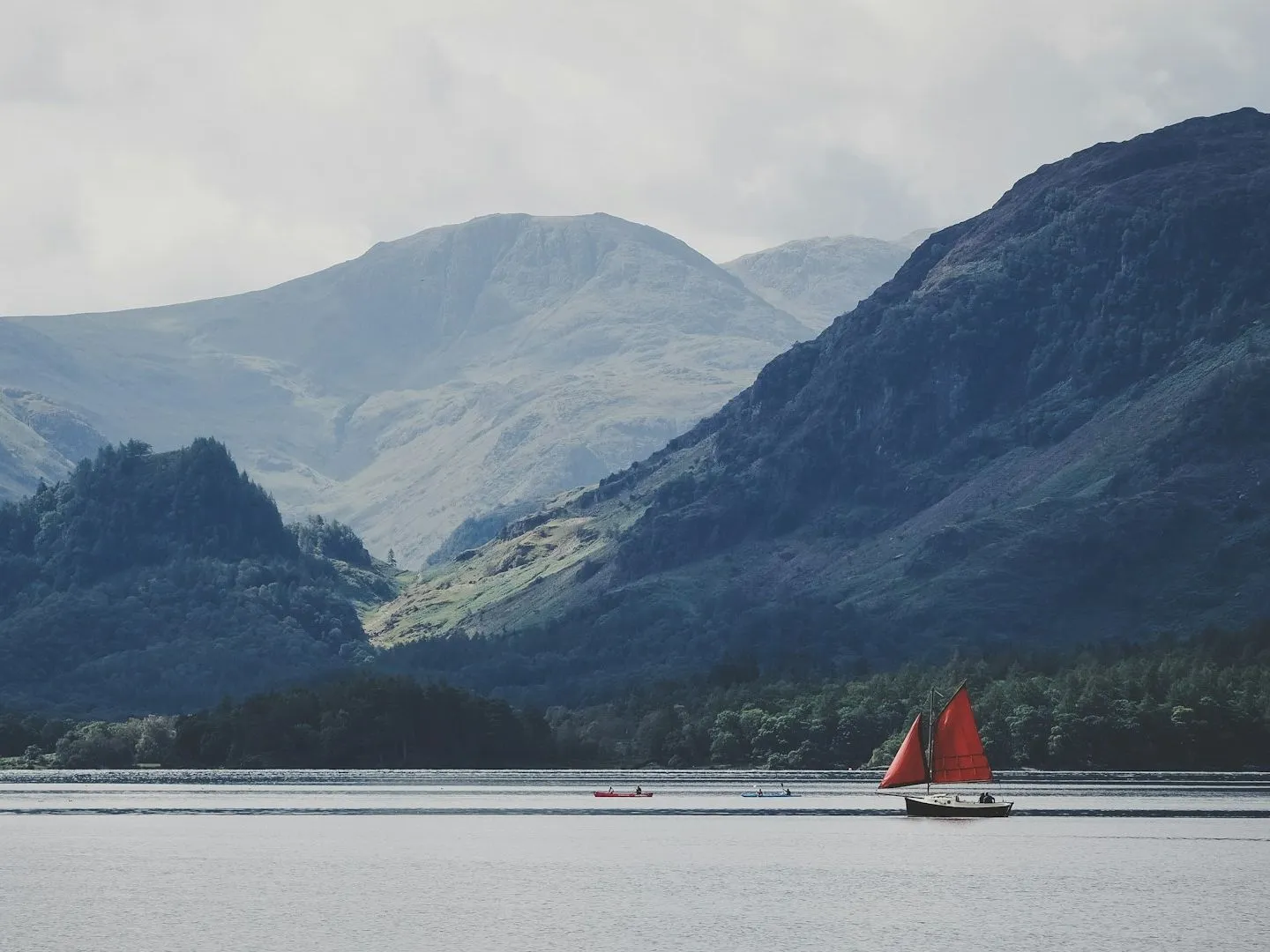 Sailing boat with bright red sails on Derwentwater beneath misty Lake District fells.