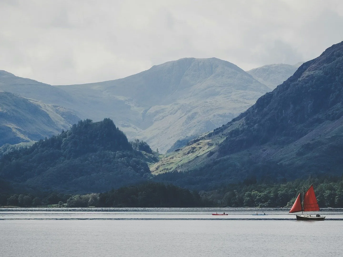 Sailing boat with bright red sails on Derwentwater beneath misty Lake District fells.