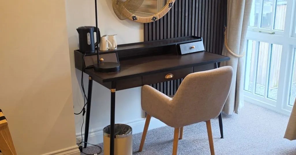 Stylish hotel room workspace with black desk, upholstered chair, round woven mirror and dark slatted feature wall.