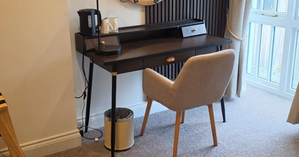 Stylish hotel room workspace with black desk, upholstered chair, round woven mirror and dark slatted feature wall.