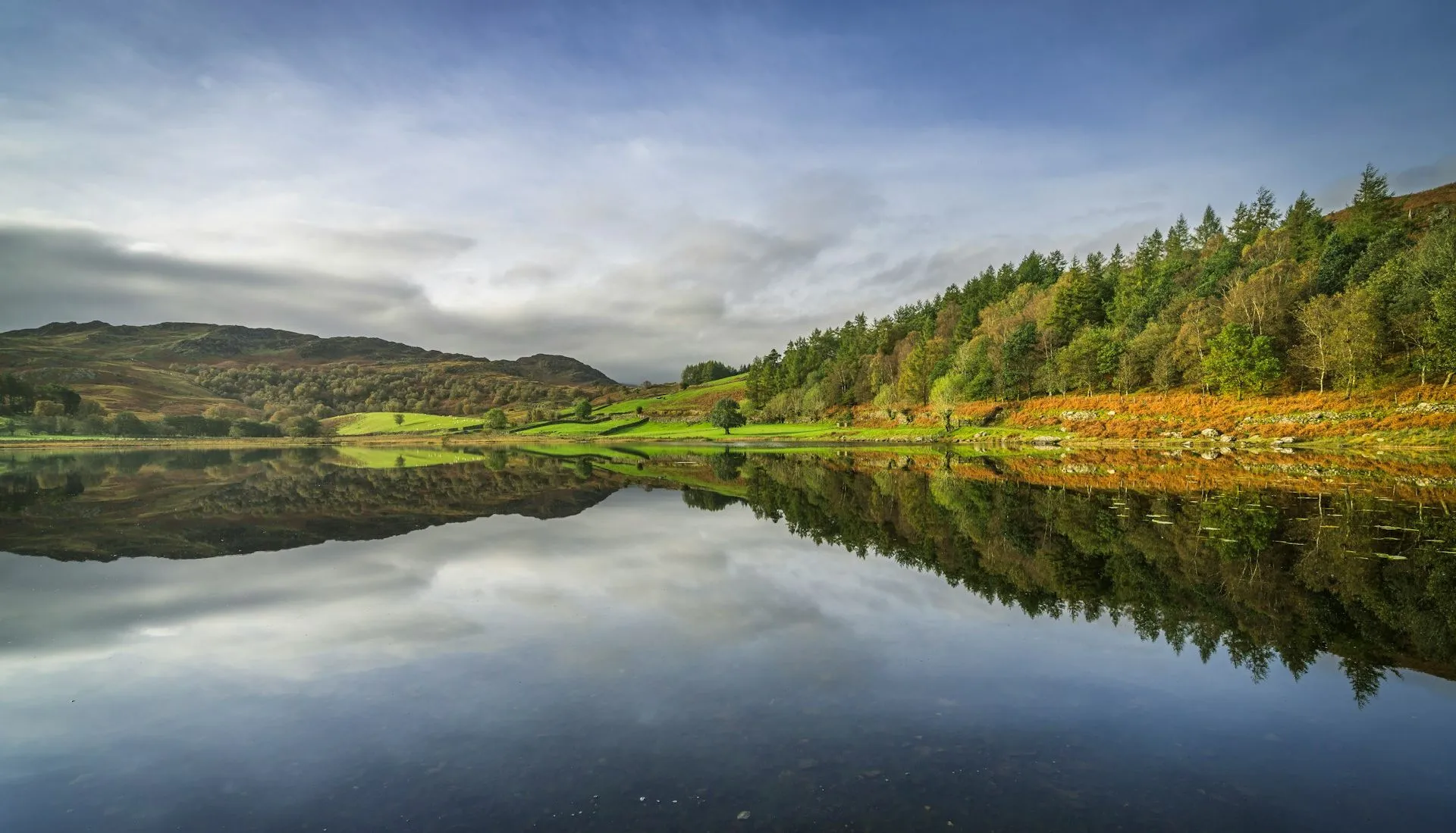Calm lake at Watendlath reflecting wooded hills, green fields and a cloudy blue sky.