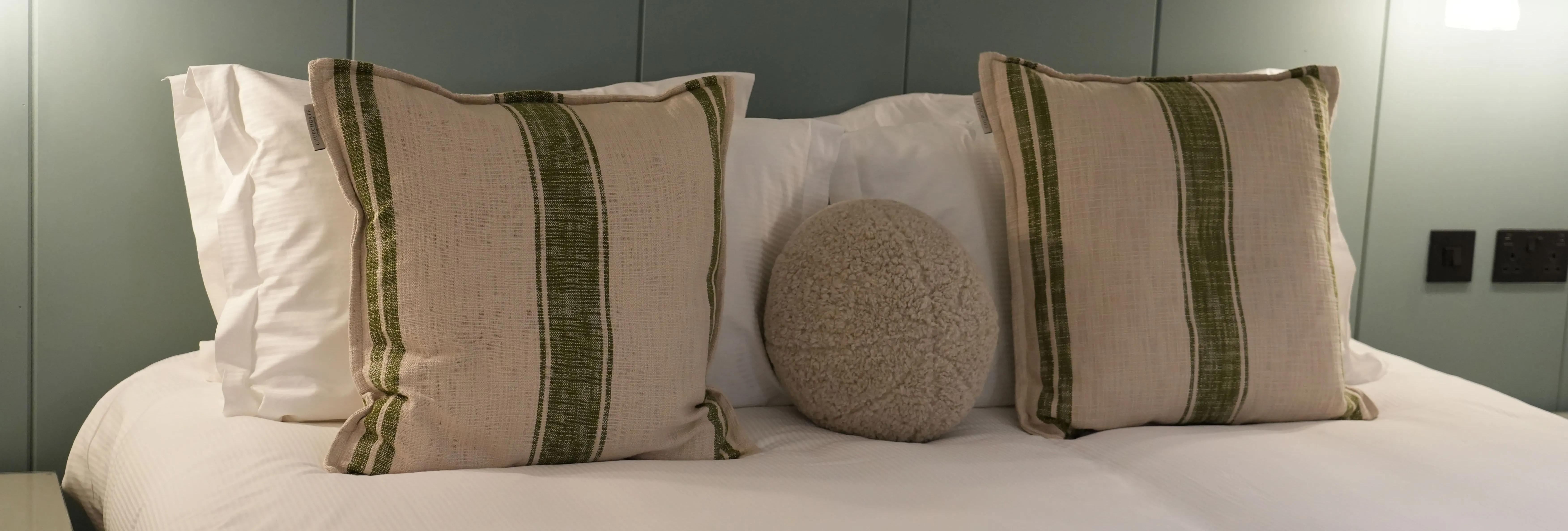 Neatly made bed with striped cushions and a round textured pillow against a muted green panelled wall