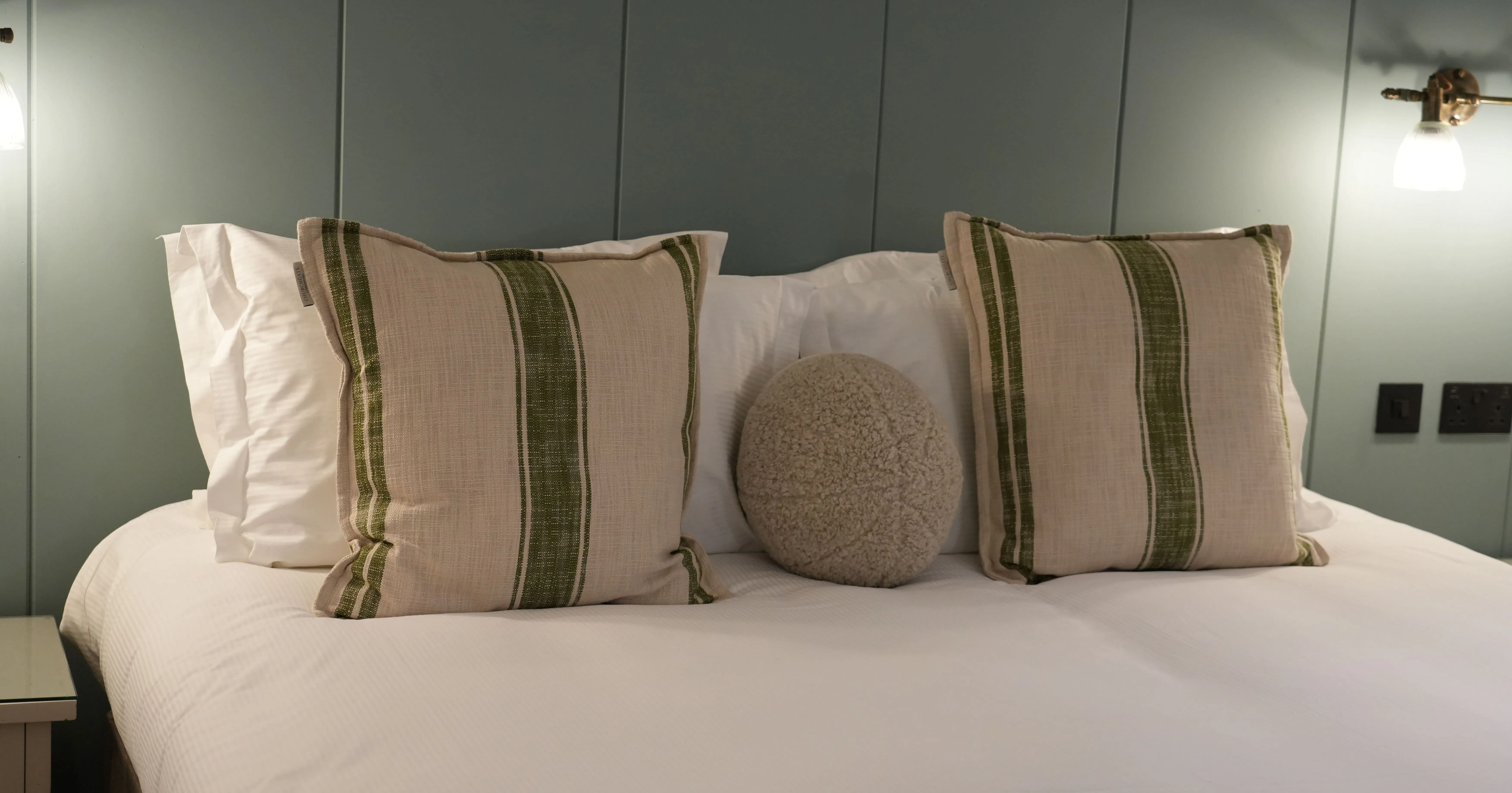 Neatly made bed with striped cushions and a round textured pillow against a muted green panelled wall