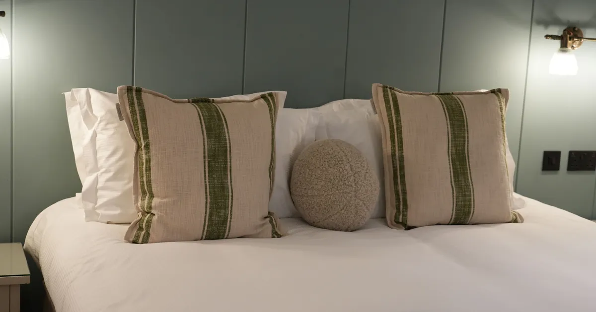 Neatly made bed with striped cushions and a round textured pillow against a muted green panelled wall