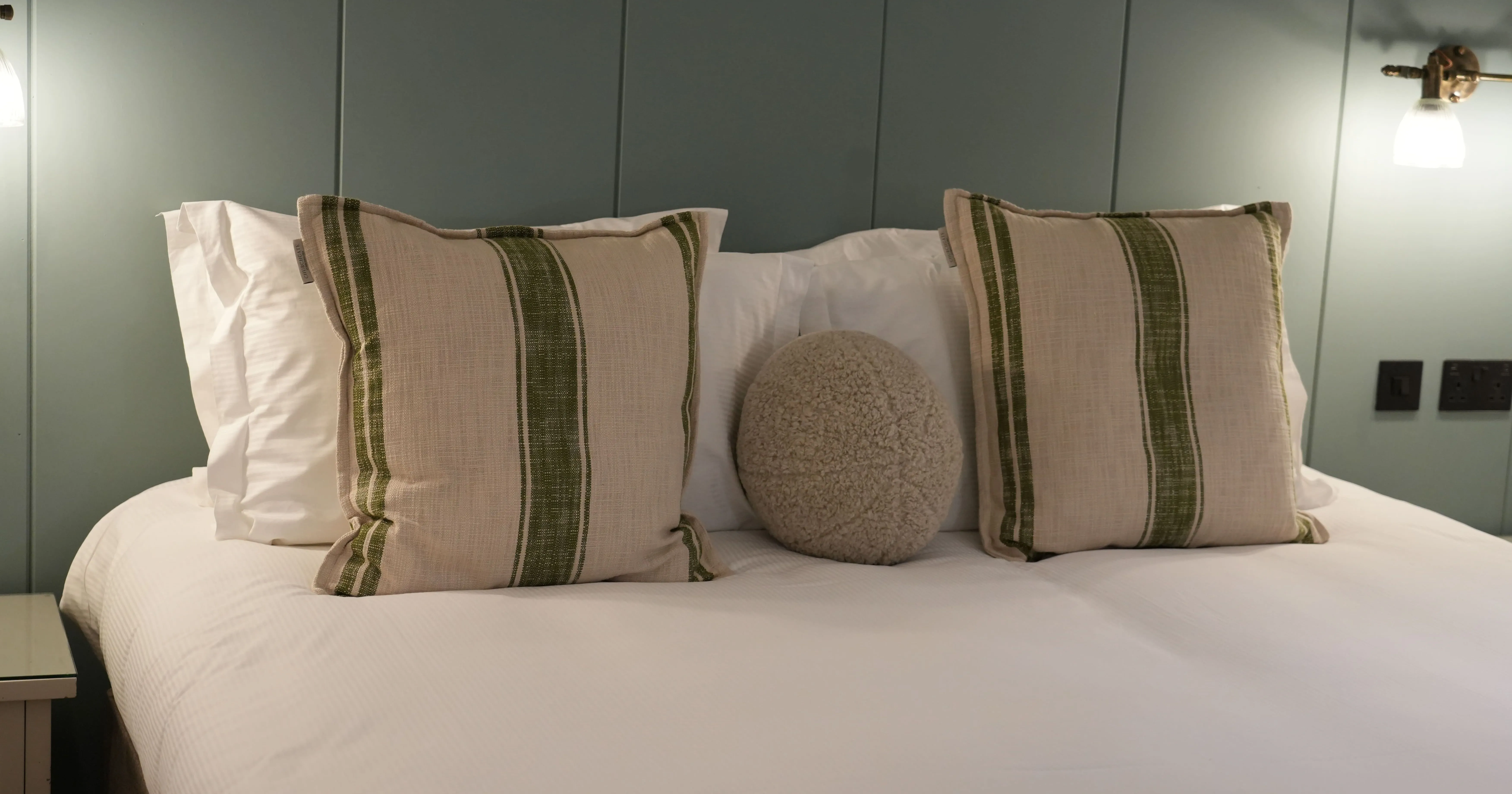 Neatly made bed with striped cushions and a round textured pillow against a muted green panelled wall
