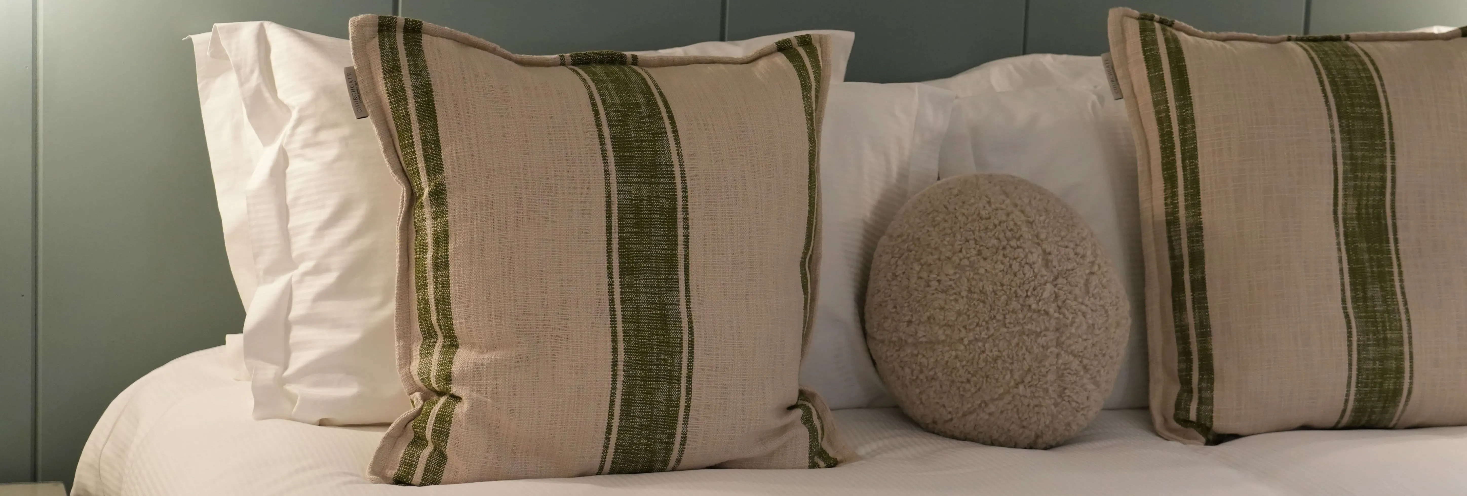 Neatly made bed with striped cushions and a round textured pillow against a muted green panelled wall