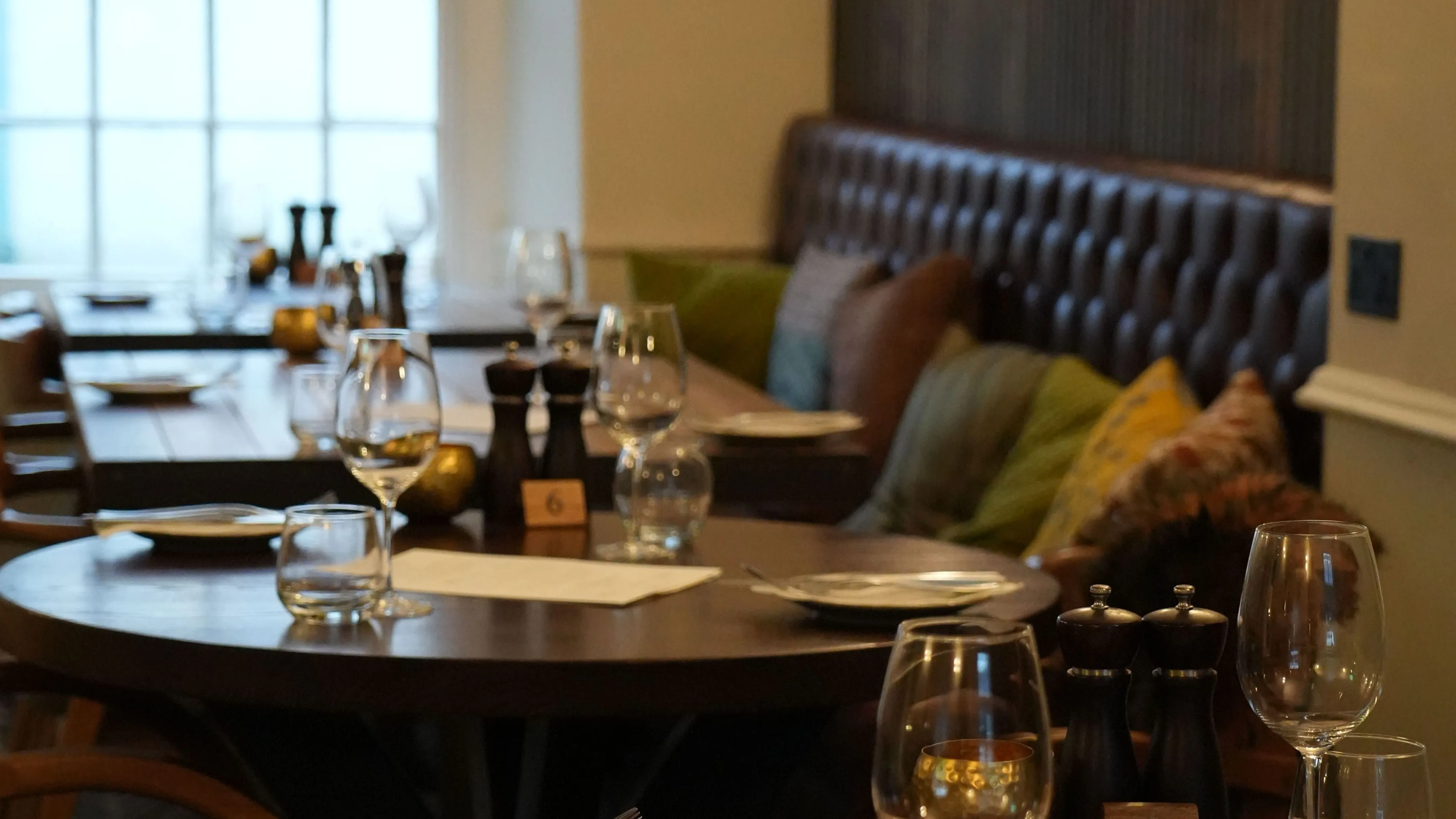 Warmly lit restaurant interior with set wooden tables, wine glasses and cushioned booth seating.