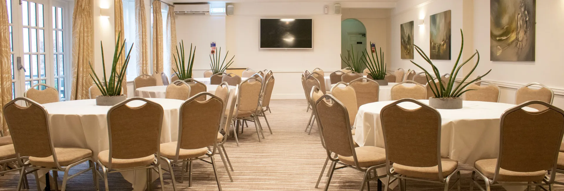 A conference room with round tables and beige chairs arranged neatly, decorated with potted plants.