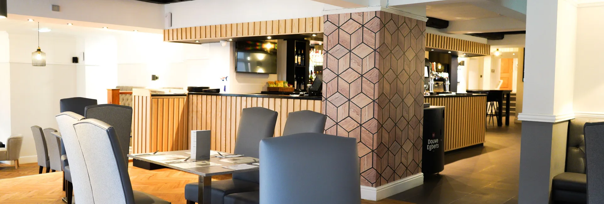 Modern restaurant interior with wooden flooring, grey chairs, and a geometric accent wall.