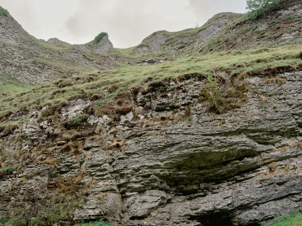 A child in a yellow jacket stands near the entrance of a rocky cave at the base of a grassy hill.
