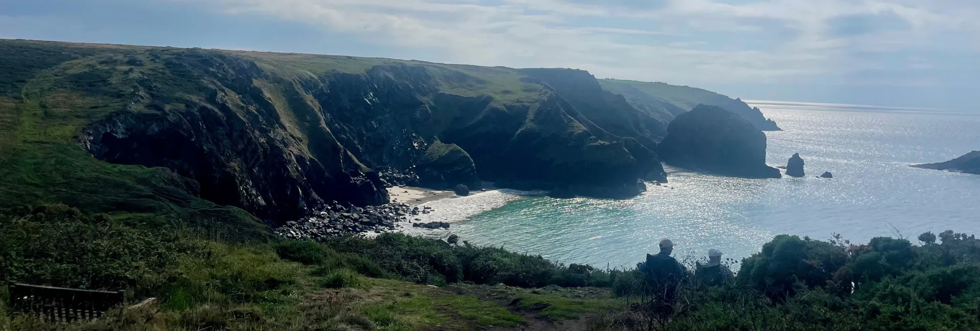 Coastal view with green cliffs, a rocky beach, and sunlight reflecting on the sea.