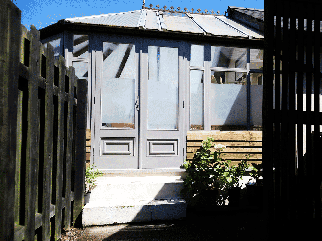 Modern garden room or conservatory with frosted glass doors, surrounded by wooden fencing and potted plants.