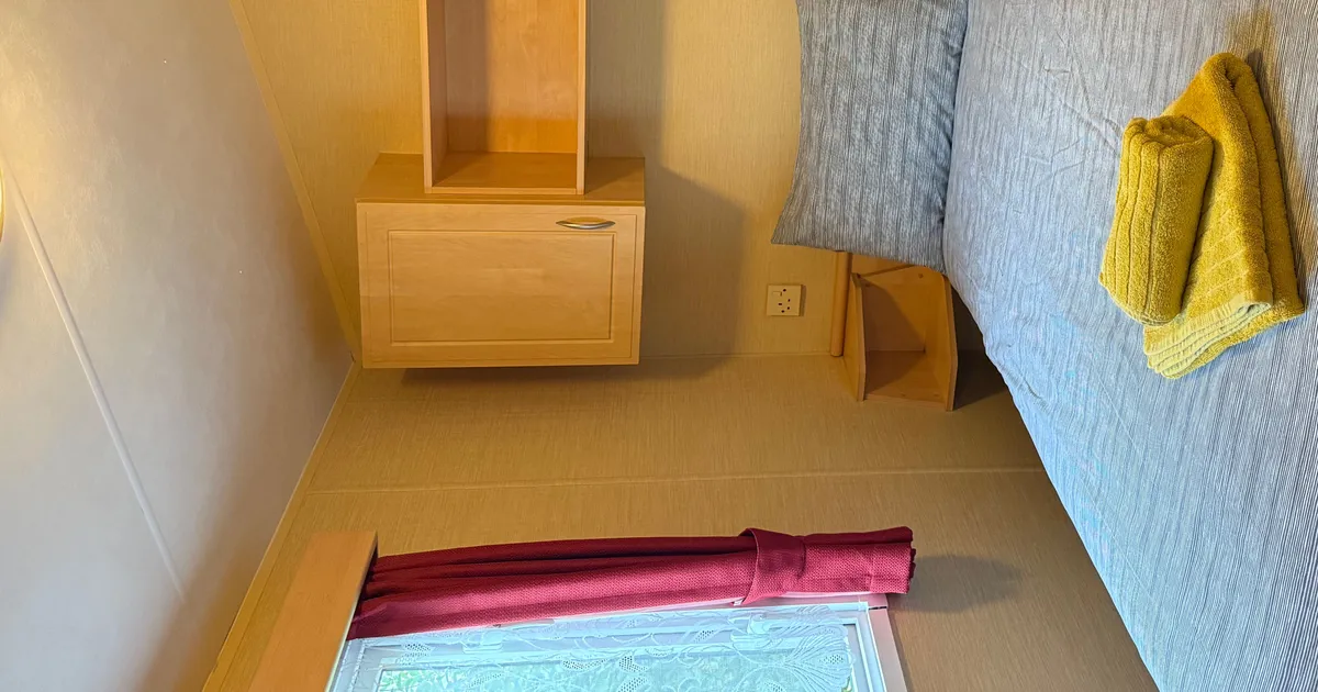 Small bedroom with a double bed, grey bedding, yellow towels, wooden cabinets, and a window with red curtains.