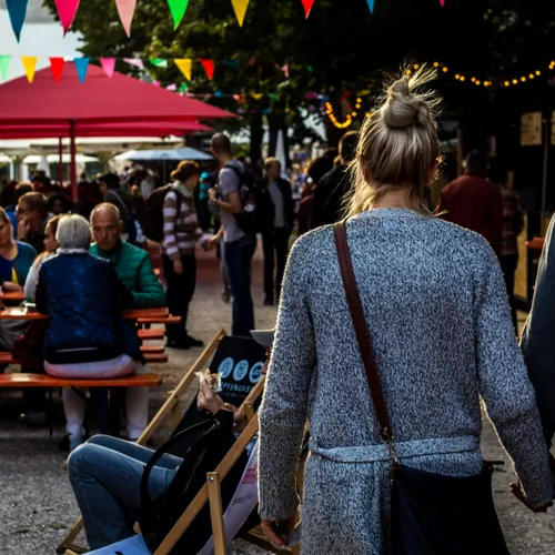 Couple holding hands walking through a lively outdoor market or festival with colorful bunting and people sitting at picnic tables.