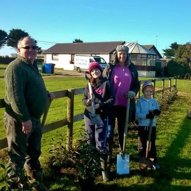 A group of people standing outdoors by a fence, planting small trees and holding gardening tools.