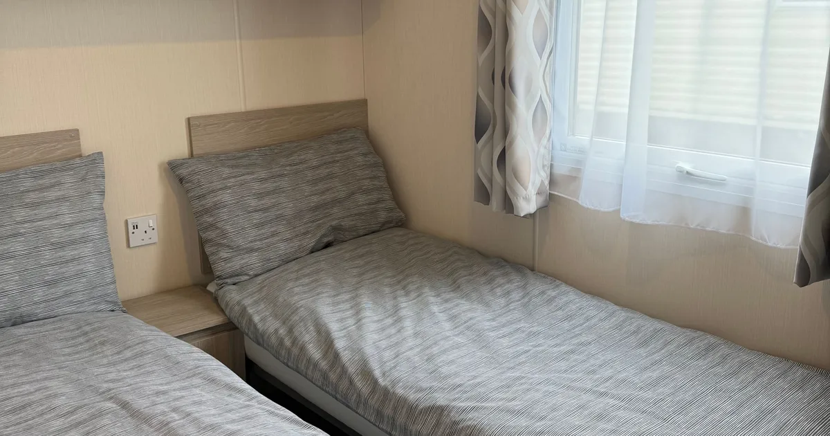 Small bedroom with two single beds, grey striped bedding, a window with patterned curtains, and wooden shelving above.