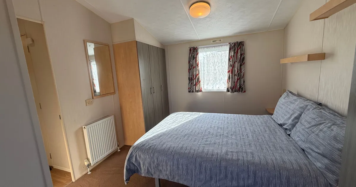 A tidy bedroom with a double bed, wardrobe, mirror, and a window with patterned curtains.