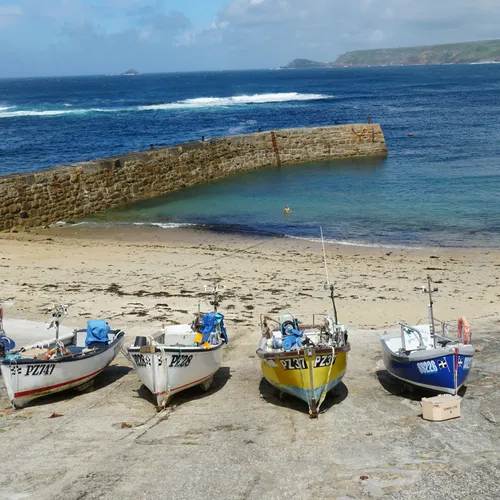 Small fishing boats lined up on a slipway near a sandy beach and a stone harbour wall with the sea in the background.